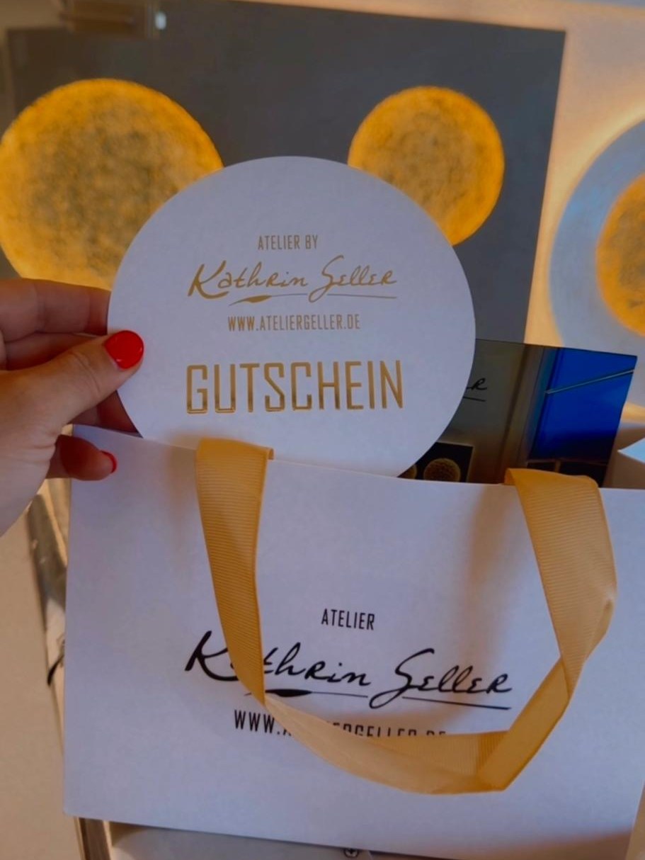 Runder Gutschein des Ateliers Kathrin Geller in einer Hand, dahinter eine Geschenktasche und beleuchtete Monde